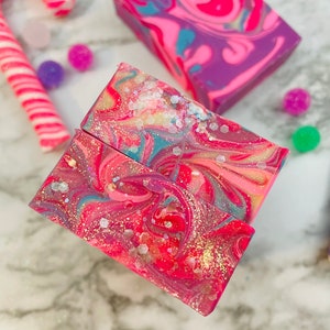 May include: Close-up of colorful handmade soap bars with a swirled design in shades of pink, purple, and blue, with glitter accents. A candy cane and small colorful balls are in the background. The soap bars are a festive, decorative item.