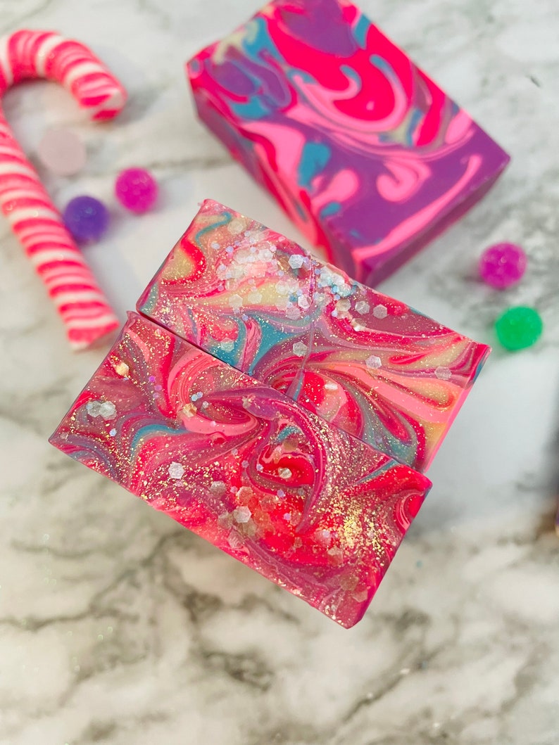 May include: Close-up of colorful handmade soap bars with a swirled design in shades of pink, purple, and blue, with glitter accents. A candy cane and small colorful balls are in the background. The soap bars are a festive, decorative item.