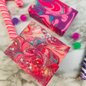 May include: Artisan soaps with vibrant swirls of pink, purple, and teal, accented with gold glitter. A pink and white striped candy cane and colorful pom-poms add a festive touch. The soaps have a marbled effect, creating a unique and decorative appearance.