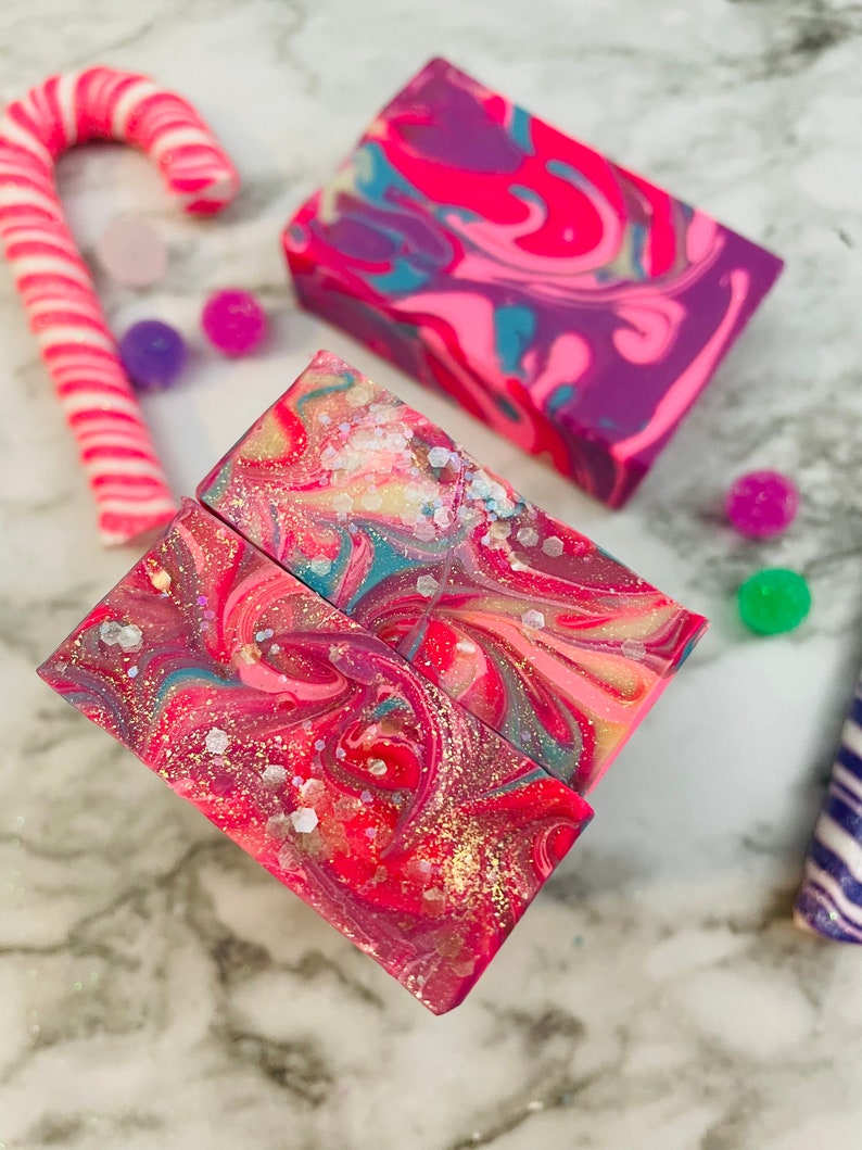 May include: Artisan soaps with vibrant swirls of pink, purple, and teal, accented with gold glitter. A pink and white striped candy cane and colorful pom-poms add a festive touch. The soaps have a marbled effect, creating a unique and decorative appearance.