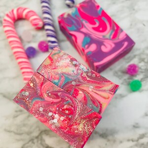 May include: A collection of handmade soaps and candy canes. The soaps feature vibrant swirls of pink, purple, and blue with glitter accents. Two pink and white candy canes and a purple and white candy cane are also present, along with colorful round candies.