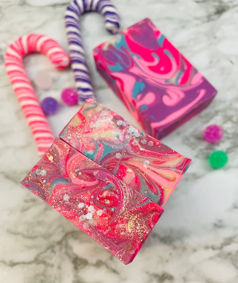 May include: A collection of handmade soaps and candy canes. The soaps feature vibrant swirls of pink, purple, and blue with glitter accents. Two pink and white candy canes and a purple and white candy cane are also present, along with colorful round candies.