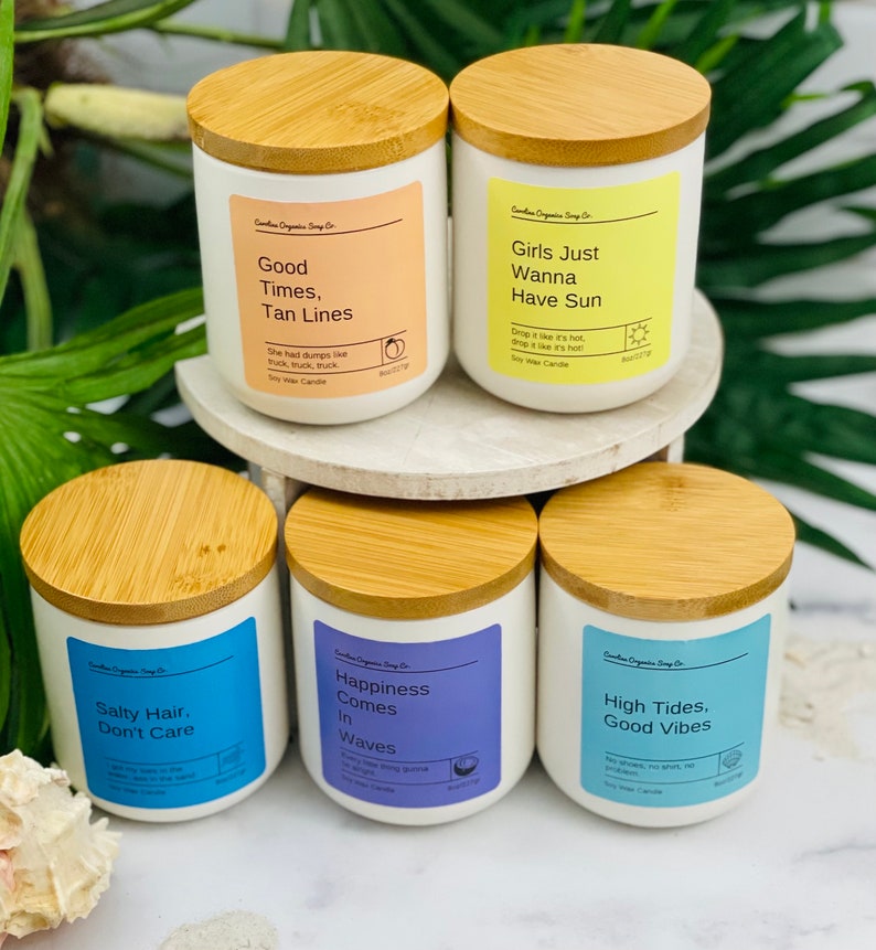 Puede incluir: Seis velas de cera de soja con tapas de madera y etiquetas coloridas. Las etiquetas presentan frases con tem&aacute;tica veraniega como "Good Times, Tan Lines", "Girls Just Wanna Have Sun", "Salty Hair, Don't Care", "Happiness Comes In Waves", y "High Tides, Good Vibes".