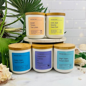 Puede incluir: Cuatro velas de cera de soja blancas con tapas de madera y etiquetas. Las etiquetas tienen diferentes citas y colores con tem&aacute;tica veraniega. Las velas est&aacute;n dispuestas en dos filas de dos sobre una superficie blanca.