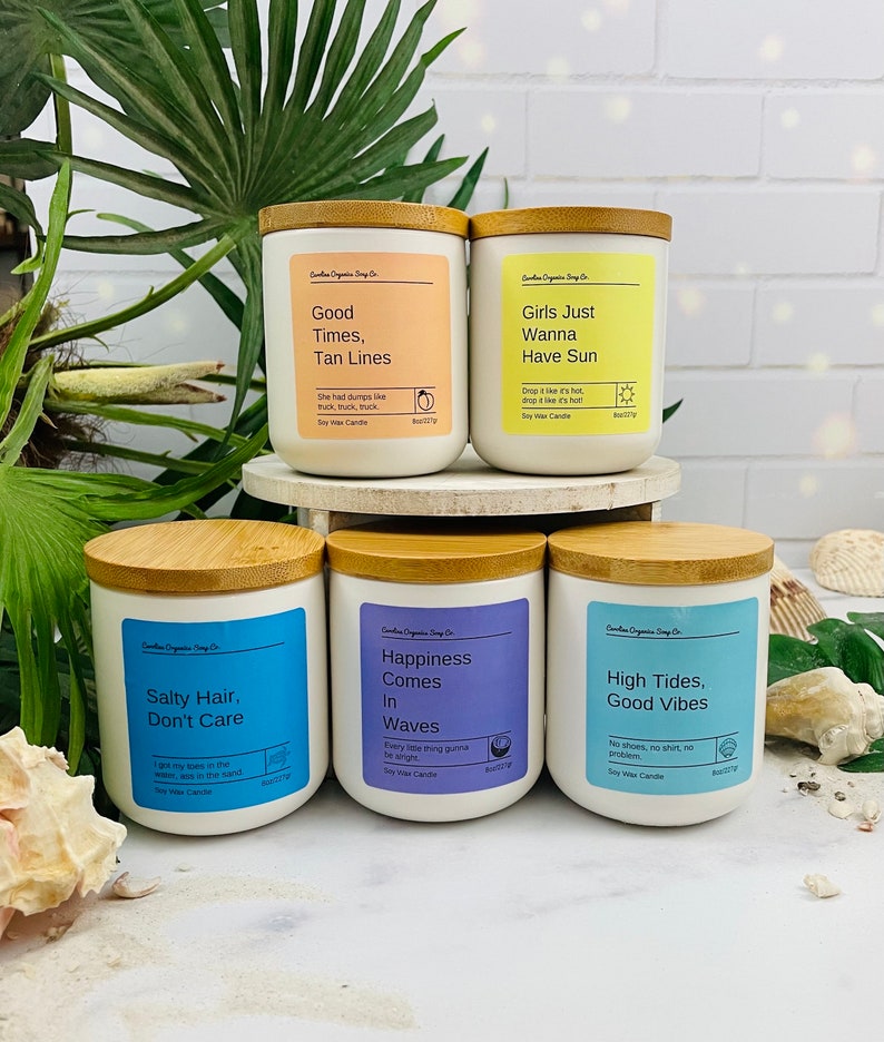 Puede incluir: Cuatro velas de cera de soja blancas con tapas de madera y etiquetas. Las etiquetas tienen diferentes citas y colores con tem&aacute;tica veraniega. Las velas est&aacute;n dispuestas en dos filas de dos sobre una superficie blanca.