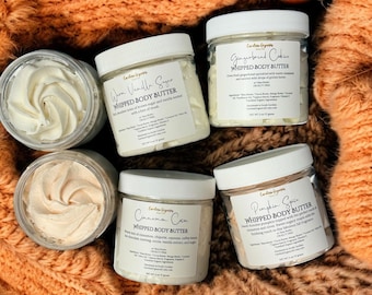May include: Six jars of whipped body butter with white lids are arranged on an orange textured surface. The labels read "Warm Vanilla Sugar," "Gingerbread Cookies," "Cinnamon Cocoa," and "Pumpkin Spice." The image is taken from above, showing the jars' contents.