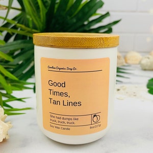 Puede incluir: Vela de cera de soja blanca con tapa de bamb&uacute;. La etiqueta es de color melocot&oacute;n con texto negro que dice "Carolina Organics Soap Co. Good Times, Tan Lines. She had dumps like truck, truck, truck. Soy Wax Candle 8oz/227gr".