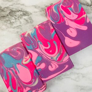 May include: Three rectangular bars of handmade soap with a swirled design. The soap features a vibrant color palette of pink, purple, and teal, creating a marbled effect. The soap bars are arranged on a white and gray marble surface.