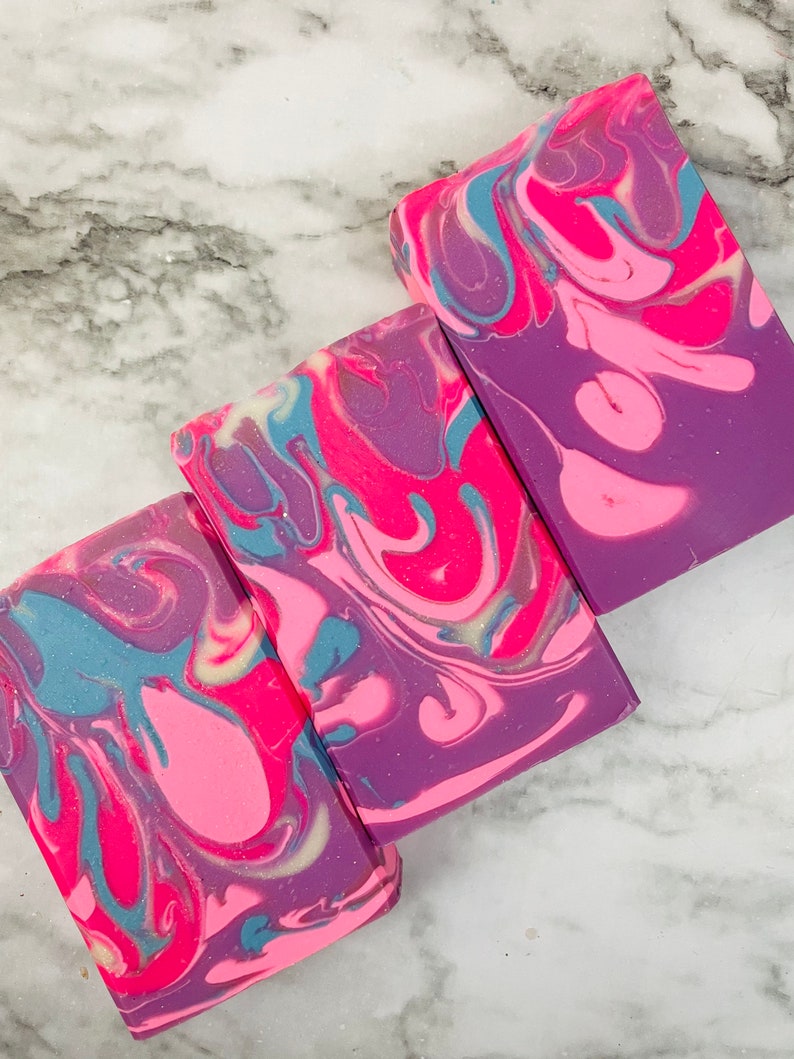 May include: Three rectangular bars of handmade soap with a swirled design. The soap features a vibrant color palette of pink, purple, and teal, creating a marbled effect. The soap bars are arranged on a white and gray marble surface.