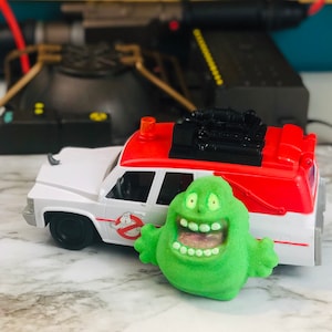 Puede incluir: Una bomba de baño verde con forma de fantasma de la película Ghostbusters. La bomba de baño tiene una sonrisa de dientes y una lengua rosa brillante. La bomba de baño está delante de un coche de juguete blanco con techo rojo y un logotipo de Ghostbusters negro en el lateral.