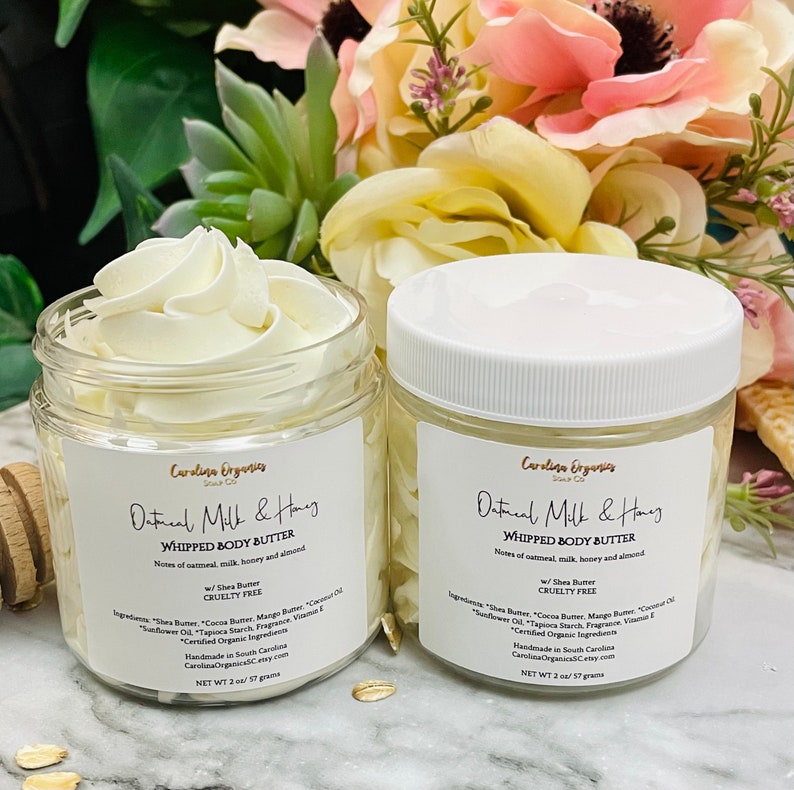 May include: Two glass jars of oatmeal milk and honey whipped body butter with white lids. The jars are labeled "Oatmeal Milk & Honey Whipped Body Butter" with the ingredients listed below. The jars are sitting on a white surface with oats scattered around them.
