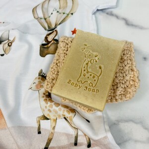 Puede incluir: Una pastilla de jab&oacute;n para beb&eacute; de color marr&oacute;n claro con un dise&ntilde;o de jirafa y las palabras "Baby Soap" est&aacute; sobre un jabonera de ganchillo beige. El jab&oacute;n y el jabonera est&aacute;n sobre un body blanco para beb&eacute; con un dise&ntilde;o de jirafa y globo aerost&aacute;tico.