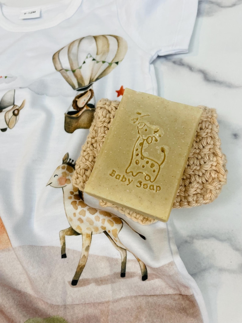 Puede incluir: Una pastilla de jab&oacute;n para beb&eacute; de color marr&oacute;n claro con un dise&ntilde;o de jirafa y las palabras "Baby Soap" est&aacute; sobre un jabonera de ganchillo beige. El jab&oacute;n y el jabonera est&aacute;n sobre un body blanco para beb&eacute; con un dise&ntilde;o de jirafa y globo aerost&aacute;tico.