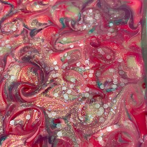 May include: Abstract art piece with a swirling pattern of pink, red, teal, and white. The design features glitter accents, creating a shimmering effect. The colors blend together in a fluid, marbled style, suggesting a handmade or artistic creation.