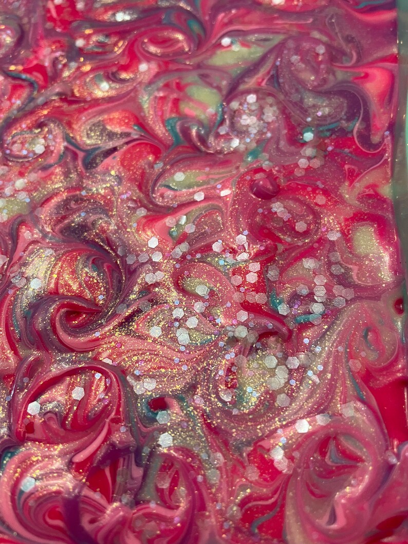 May include: Abstract art piece with a swirling pattern of pink, red, teal, and white. The design features glitter accents, creating a shimmering effect. The colors blend together in a fluid, marbled style, suggesting a handmade or artistic creation.