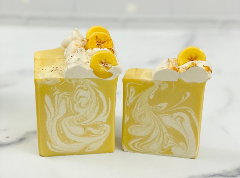 K&ouml;nnte beinhalten: Zwei rechteckige Seifenst&uuml;cke mit einem gelb-wei&szlig;en Wirbelmuster. Die Oberseite jeder Seife ist mit wei&szlig;em Zuckerguss, Bananenscheiben und einer Best&auml;ubung mit braunem Pulver verziert. Die Seifen haben ein Dessert-Thema.