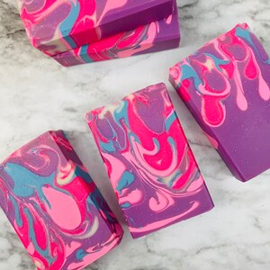 May include: Rectangular bars of handmade soap in shades of purple, pink, and blue. The soap features a swirled design with a marbled effect. The bars are arranged on a white and gray marble surface.