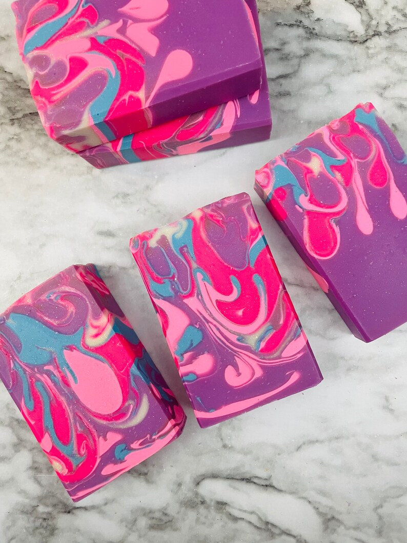 May include: Rectangular bars of handmade soap in shades of purple, pink, and blue. The soap features a swirled design with a marbled effect. The bars are arranged on a white and gray marble surface.