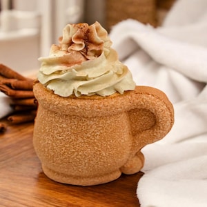 Puede incluir: Una taza de cerámica marrón con forma de taza de café con una cobertura de crema batida blanca y una pizca de canela encima.