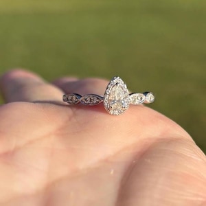 May include: A silver-toned ring featuring a central pear-shaped stone surrounded by a halo of small round stones. The band is detailed with a series of marquise-shaped metal settings, each containing tiny embedded stones, extending from the center.