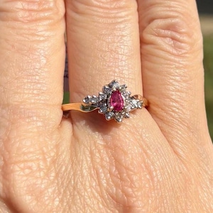 May include: A gold ring featuring a central pear-shaped pink gemstone surrounded by a cluster of small, clear, round-cut stones. The setting is crafted from a metallic band that holds the central pink stone and the surrounding clear stones securely.