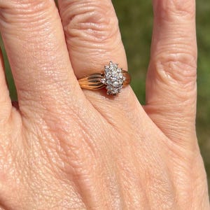 May include: A gold ring featuring a cluster of small, clear, round-cut stones arranged in an oval shape. The band of the ring is smooth and polished, with a tapered design that widens slightly as it reaches the central stone setting.