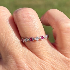 May include: A silver-toned ring featuring an alternating pattern of round red stones and small clear stones set within marquise and diamond-shaped metal frames. The band has a delicate, textured milgrain edge detail surrounding each of the stones.