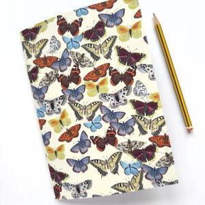 Pode incluir: Um caderno com uma capa creme adornada com um padrão de borboletas coloridas em tons de laranja, azul, amarelo e castanho. Um lápis listrado amarelo e preto está sobre o fundo branco ao lado do caderno.