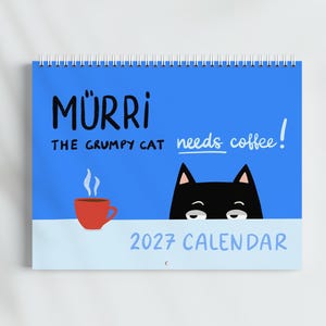 Puede incluir: Un calendario de pared 2027 con fondo azul brillante que muestra un gato negro asomándose por un borde azul claro. El texto dice MÜRRI THE GRUMPY CAT needs coffee! Una taza de café roja con vapor está junto al gato. Encuadernación en espiral.