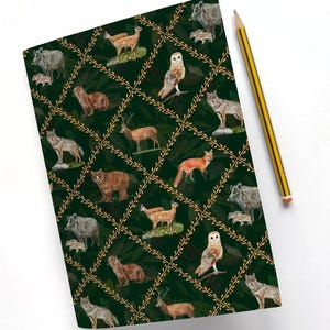 May include: A notebook featuring a dark green cover with a repeating pattern of woodland creatures such as deer, owls, and foxes. The animals are set in a grid with gold-coloured vines. A yellow and black pencil lies on a white surface.
