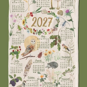 May include: A cream-coloured tea towel featuring a 2027 calendar with botanical and wildlife illustrations. The design includes months from January to December, surrounded by watercolour drawings of mushrooms, insects, birds, a hedgehog, and various plants.
