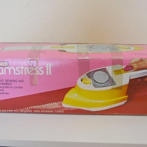 May include: A pink box for a Steamstress II sewing aid. The box features a yellow and white iron design, with the text "Steamstress II" and "The Greatest Sewing Aid Since The Thimble".