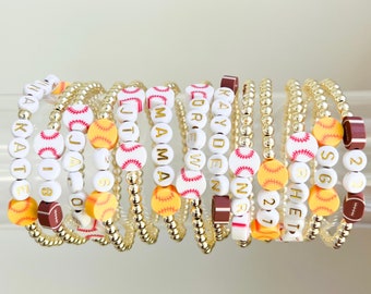 May include: A collection of beaded bracelets with white letter beads spelling out names and numbers. The bracelets feature gold-colored beads and sports-themed accents, including baseballs and footballs. The bracelets are designed for a sporty theme.