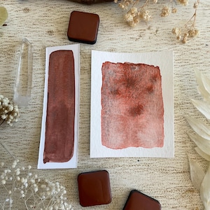 May include: An overhead shot features watercolor art supplies and decorative elements. Two square, dark brown paint containers are positioned near two watercolor paper swatches. One swatch is rectangular, the other square. A clear crystal and dried flowers are also present.