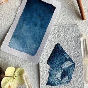Peut inclure: Deux peintures à l'aquarelle sur papier texturé. L'une présente un dégradé bleu rectangulaire, l'autre une forme abstraite bleue. Des bâtons de cannelle, des cristaux et une fleur séchée sont disposés autour.