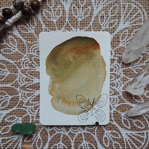 Op de afbeelding: Een aquarel schilderij van een vlinder op een witte kaart met een bruine en groene aquarel was in de achtergrond. De kaart ligt op een jute oppervlak met een wit mandala ontwerp.