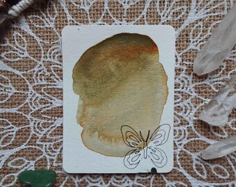 May include: A watercolor painting of a butterfly on a white card with a brown and green watercolor wash in the background. The card is on a burlap surface with a white mandala design.