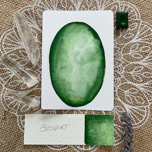 Peut inclure: Une peinture à l'aquarelle d'une forme ovale verte avec le mot "BOSQUET" écrit sur une carte blanche en dessous. Un petit pot de peinture verte est également visible.