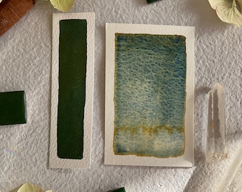 May include: An artistic composition featuring watercolor paintings and green square tiles. The paintings showcase shades of green and blue, with a textured appearance. Dried flowers and a clear crystal are also present, creating a natural aesthetic.
