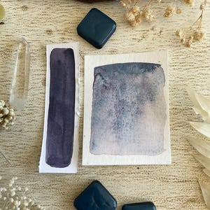 Peut inclure: Vue aérienne de peintures aquarelles et de godets de peinture carrés bleu foncé. Les échantillons d'aquarelle sont sur papier blanc, présentant des nuances d'indigo et de bleu. Des fleurs séchées et une baguette de cristal complètent la composition.