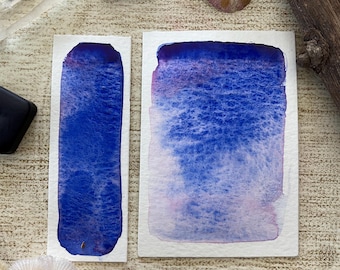 May include: Two watercolor paint swatches in shades of blue and purple on white paper. The swatches are surrounded by black square objects, dried flowers, and a clear crystal. The paint colors blend from dark to light.