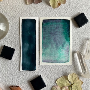 Peut inclure: Deux peintures à l'aquarelle aux teintes vert foncé et vert-bleu. Les peintures sont rectangulaires et entourées de fleurs séchées, de cristaux et de pierres carrées noires. L'œuvre est exposée sur une surface texturée blanc cassé.