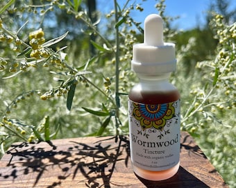 May include: A small glass bottle of Wormwood Tincture with a white dropper top. The label features a colorful mandala design and the text "Wormwood Tincture". The bottle is 1 fl oz. The background includes green foliage and a blue sky.