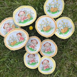 May include: A collection of daisy-themed coasters. The coasters feature illustrations of a girl and a cat in a field of daisies, with text that reads "I Love Daisies!" and "Oh How We Love Daisies!". The coasters have a yellow scalloped edge.