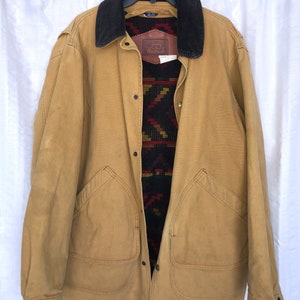 May include: A tan canvas jacket with a corduroy collar and a patterned fleece lining. The jacket has a button closure and two large pockets on the front. The label on the jacket reads "Woolrich".
