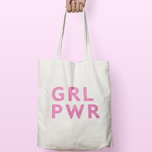 May include: A white tote bag with the pink text "GRL PWR" is held by a hand against a pink background. The bag has long straps and appears to be made of canvas or a similar fabric. The text is in a bold, sans-serif font.