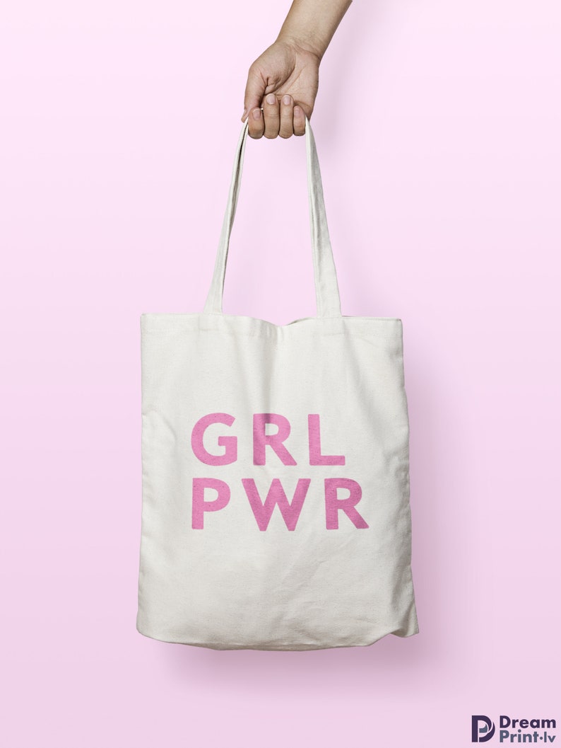 May include: A white tote bag with the pink text "GRL PWR" is held by a hand against a pink background. The bag has long straps and appears to be made of canvas or a similar fabric. The text is in a bold, sans-serif font.