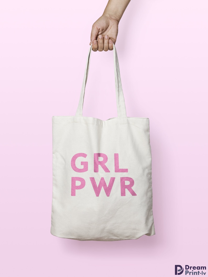 May include: A white canvas tote bag with the pink text "GRL PWR" is held by a hand against a pink background. The bag has long straps and is made of a sturdy material, suitable for carrying various items.