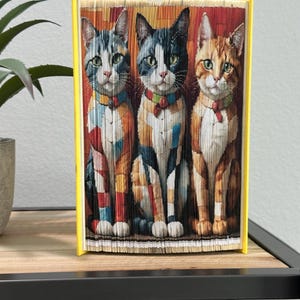 May include: A decorative book with a yellow spine featuring a folded paper art design of three cats. The cats have colorful patterned fur and are wearing bow ties. The book is displayed on a wooden shelf.