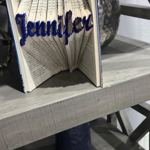 May include: A book with pages folded to spell the name "Jennifer" in blue. The book is open, revealing the folded pages that form the letters. The name is in a cursive font. The book is on a gray shelf with a decorative sphere in the background.
